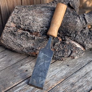 May include: A woodworking chisel with a wooden handle and a steel blade. The chisel is resting on a weathered wooden surface, with a piece of bark in the background. The handle is light brown, and the blade is a darker gray color.