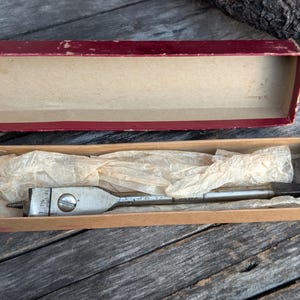 May include: A vintage wood boring bit set in its original box. The bit is silver-toned with a screw tip and measurement markings. The box is cardboard with a maroon lid and a tan interior, and is lined with tissue paper.
