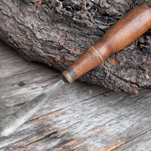 May include: A vintage wood chisel with a dark wooden handle and a metal blade. The blade is slightly curved and shows signs of wear. The chisel is resting on a weathered wooden surface, suggesting it's a well-used tool.