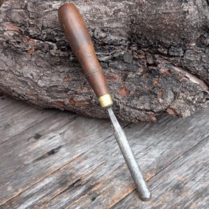 May include: A woodworking chisel with a dark wooden handle and a metal blade. The handle is a rich brown color, and the blade is a silver-gray. The chisel is designed for shaping and carving wood. The tool is set against a rustic wooden background.
