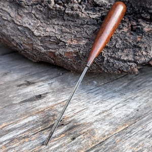 May include: A vintage awl with a dark wooden handle and a slender, silver-colored metal blade. The awl is positioned diagonally against a weathered wooden surface and a textured tree bark background.