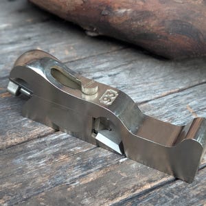 May include: A silver-colored metal hand plane tool featuring a polished finish, a sharp cutting blade, and a small adjustment knob on the top. The number 93 is clearly embossed on the side of the metal body, which has a curved, ergonomic shape.