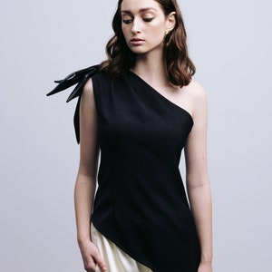May include: A woman wearing a black one-shoulder top with a black bow detail on the shoulder and white pants.