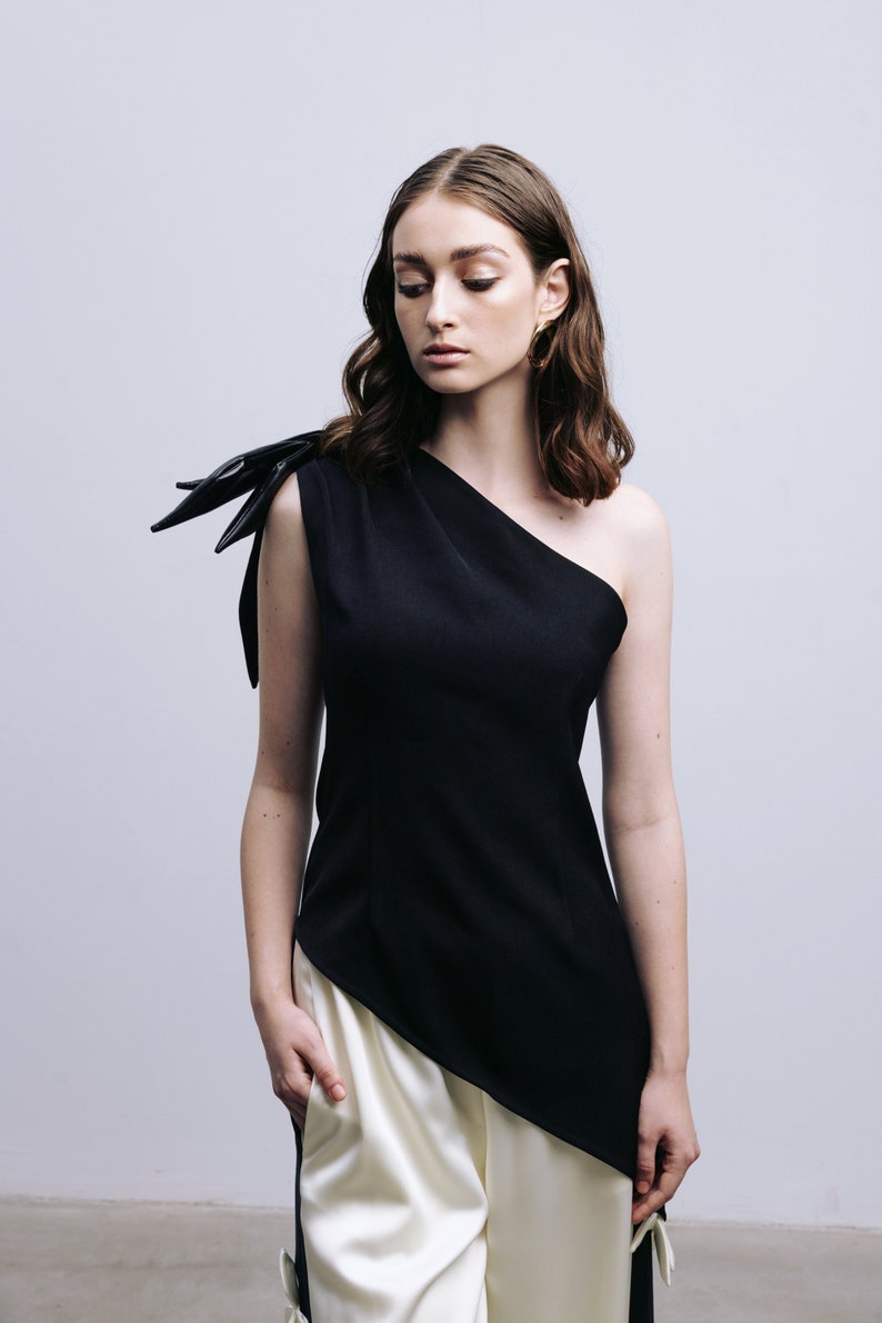 May include: A woman wearing a black one-shoulder top with a black bow detail on the shoulder and white pants.