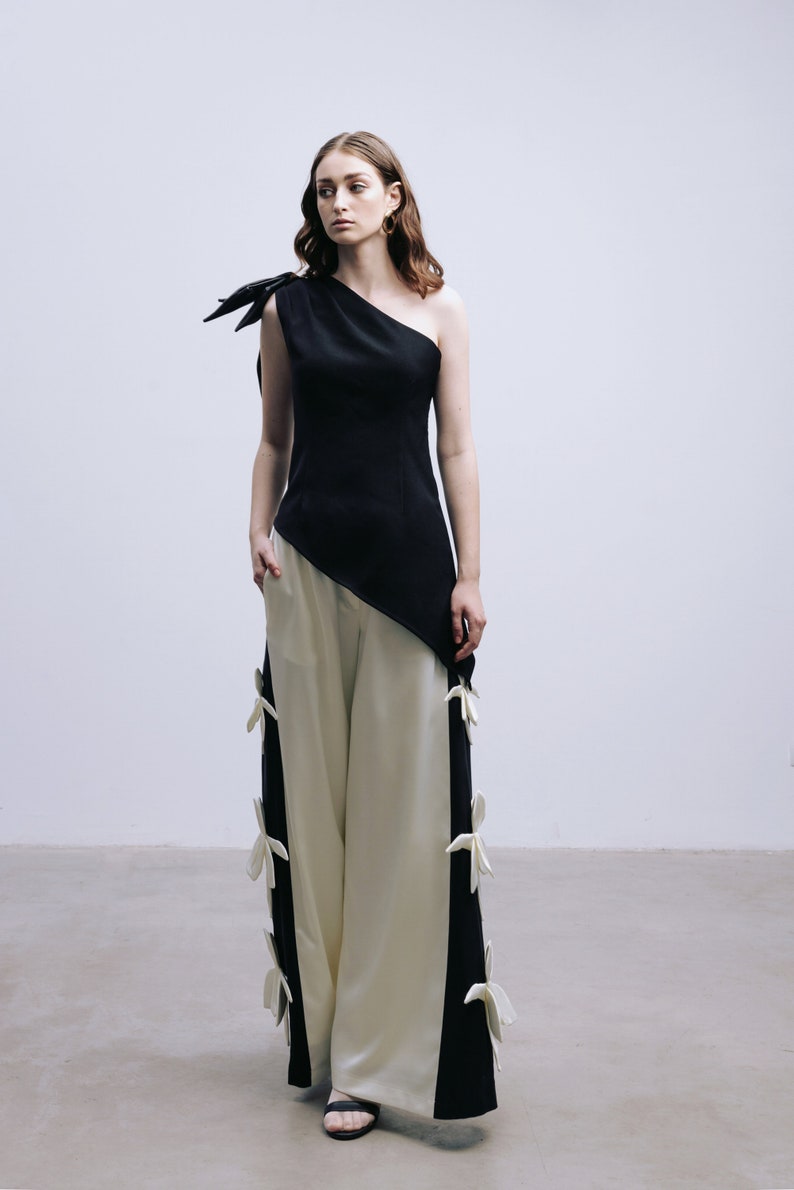 May include: A woman wearing a black one-shoulder top with a white and black asymmetrical pants. The pants have white bows on the sides.