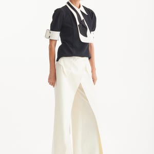 Op de afbeelding: Een zwart-witte outfit met een top met korte mouwen en contrasterende witte kraag en manchetten, gecombineerd met een witte broek met wijde pijpen en hoge taille. Zwarte schoenen.