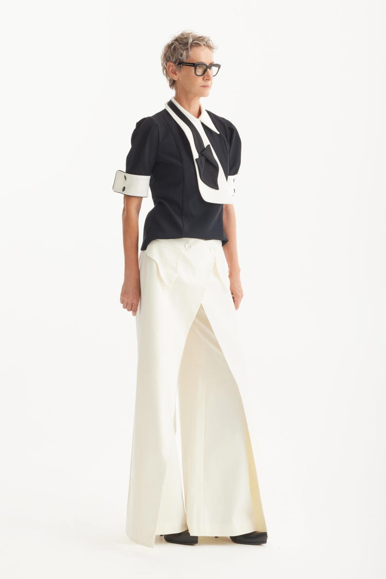 Op de afbeelding: Een zwart-witte outfit met een top met korte mouwen en contrasterende witte kraag en manchetten, gecombineerd met een witte broek met wijde pijpen en hoge taille. Zwarte schoenen.