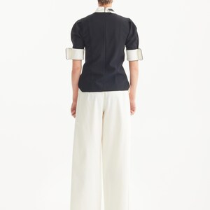 Op de afbeelding: Een zwarte top met witte kraag en manchetten, gecombineerd met een witte broek met wijde pijpen. De top heeft korte mouwen en een getailleerde pasvorm. De outfit is eenvoudig en elegant, met een minimalistische uitstraling.