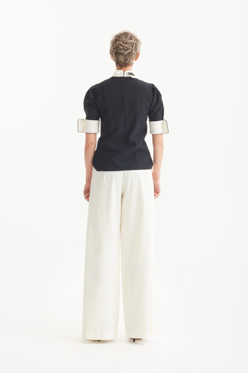 Op de afbeelding: Een zwarte top met witte kraag en manchetten, gecombineerd met een witte broek met wijde pijpen. De top heeft korte mouwen en een getailleerde pasvorm. De outfit is eenvoudig en elegant, met een minimalistische uitstraling.