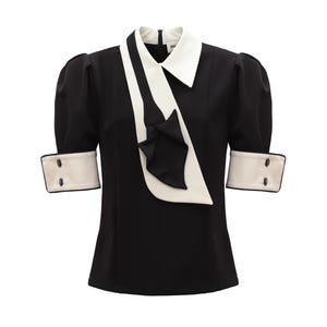 Op de afbeelding: Zwarte blouse met een witte kraag en manchetten. De top met korte mouwen heeft een wit sjaalachtig detail en een kleine strik. De manchetten hebben twee zwarte knopen. De blouse is een klassieke, elegante stijl.