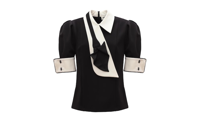 Op de afbeelding: Zwarte blouse met een witte kraag en manchetten. De top met korte mouwen heeft een wit sjaalachtig detail en een kleine strik. De manchetten hebben twee zwarte knopen. De blouse is een klassieke, elegante stijl.