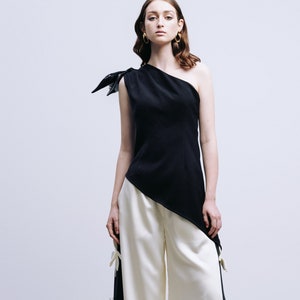 May include: A woman wearing a black one-shoulder top with a white asymmetrical hem and white wide-leg pants with white flower embellishments.