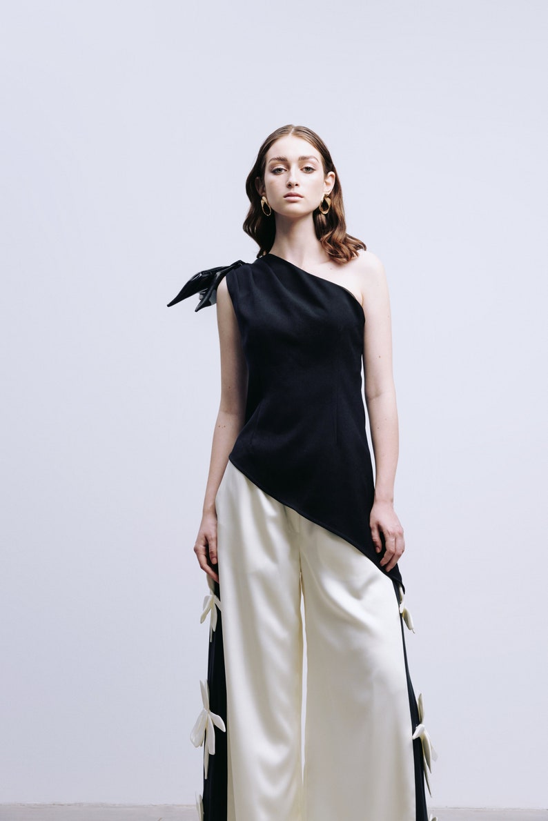 May include: A woman wearing a black one-shoulder top with a white asymmetrical hem and white wide-leg pants with white flower embellishments.