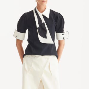 Op de afbeelding: Een zwarte blouse met witte kraag, manchetten en strikdetail. De top met korte mouwen wordt gecombineerd met een witte broek met hoge taille. De outfit is een moderne interpretatie van een klassieke stijl, geschikt voor diverse gelegenheden.