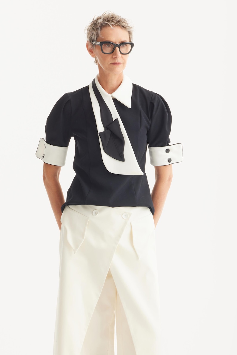 Op de afbeelding: Een zwarte blouse met witte kraag, manchetten en strikdetail. De top met korte mouwen wordt gecombineerd met een witte broek met hoge taille. De outfit is een moderne interpretatie van een klassieke stijl, geschikt voor diverse gelegenheden.