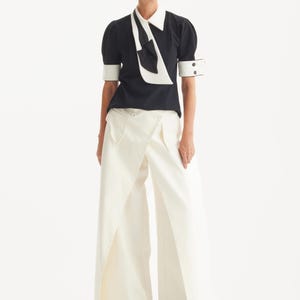 Op de afbeelding: Een monochrome outfit met een zwarte blouse met een witte kraag en strik, gecombineerd met een cr&egrave;mekleurige broek met wijde pijpen. De outfit wordt gecompleteerd met een bril met zwart montuur en schoenen, wat een chique, eigentijdse uitstraling geeft.