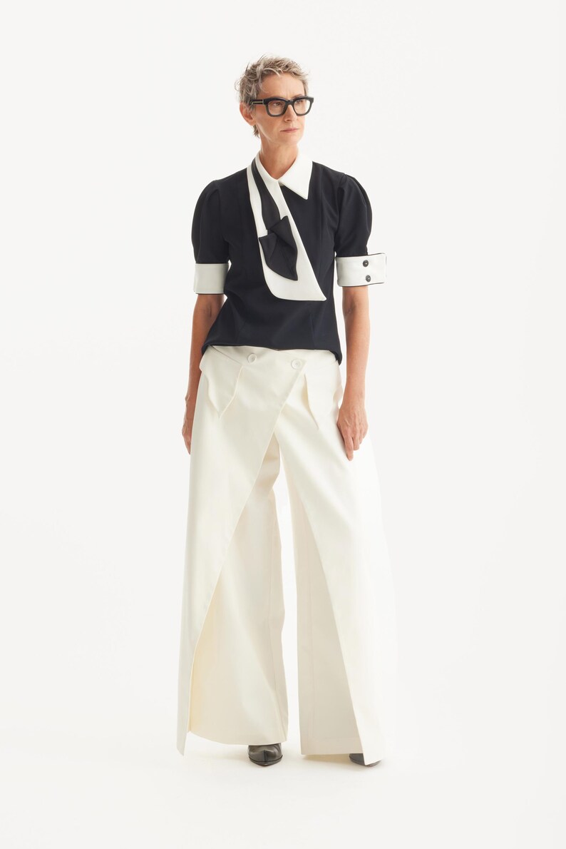 Op de afbeelding: Een monochrome outfit met een zwarte blouse met een witte kraag en strik, gecombineerd met een cr&egrave;mekleurige broek met wijde pijpen. De outfit wordt gecompleteerd met een bril met zwart montuur en schoenen, wat een chique, eigentijdse uitstraling geeft.