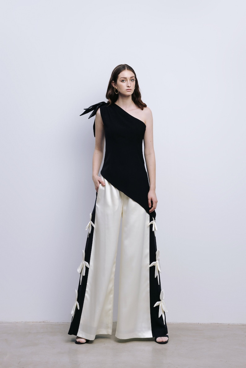 May include: A woman wearing a black one-shoulder top with a white bow detail and white wide-leg pants with black side panels and white bow details.
