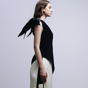 May include: A woman wearing a black sleeveless top with a black leather bow on the shoulder and white pants with black accents and white flower embellishments.
