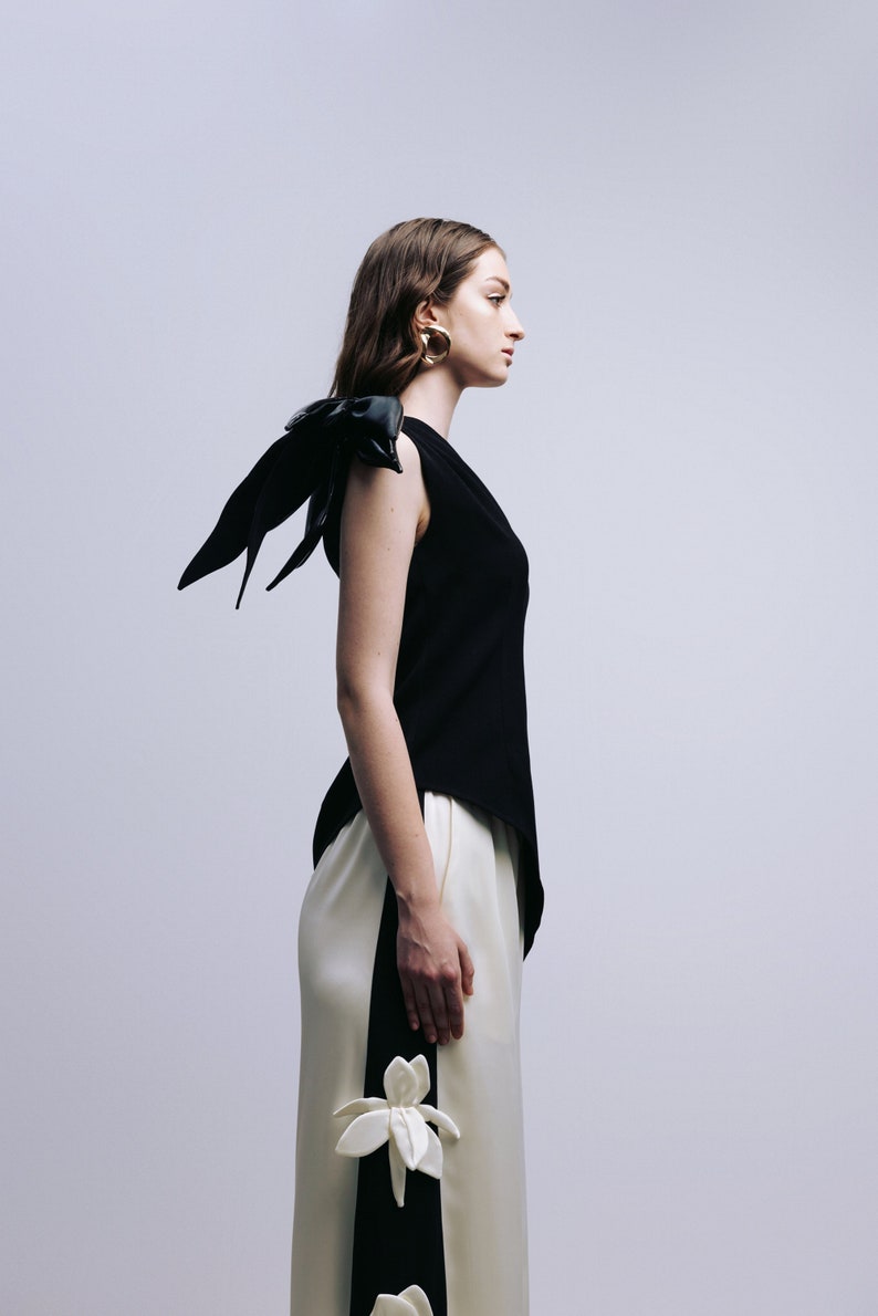 May include: A woman wearing a black sleeveless top with a black leather bow on the shoulder and white pants with black accents and white flower embellishments.
