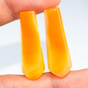 May include: Two polished, orange-yellow gemstone pendants. Each pendant has a rectangular top that tapers to a pointed bottom. The gemstones are held between fingers against a white background.