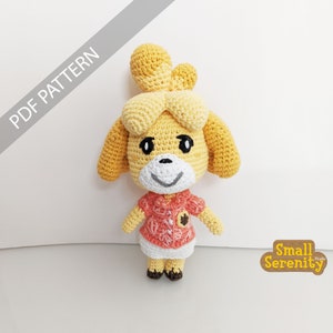 Puede incluir: Patrón de crochet para un personaje de perro amarillo y marrón que lleva una camisa rosa y blanca. El patrón se llama "Small Serenity Studio".