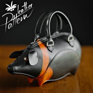 Puede incluir: Un bolso de mano de cuero negro con forma de cerdo, con rayas naranjas y orejas pequeñas. El bolso tiene un asa superior y el texto "Leather Pattern" en cursiva blanca.