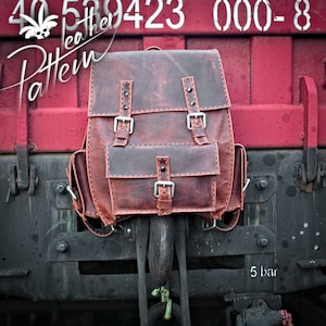 May include: A brown leather backpack with two large buckles and a smaller pocket on the front. The backpack is hanging from a metal hook on a red and black train car.
