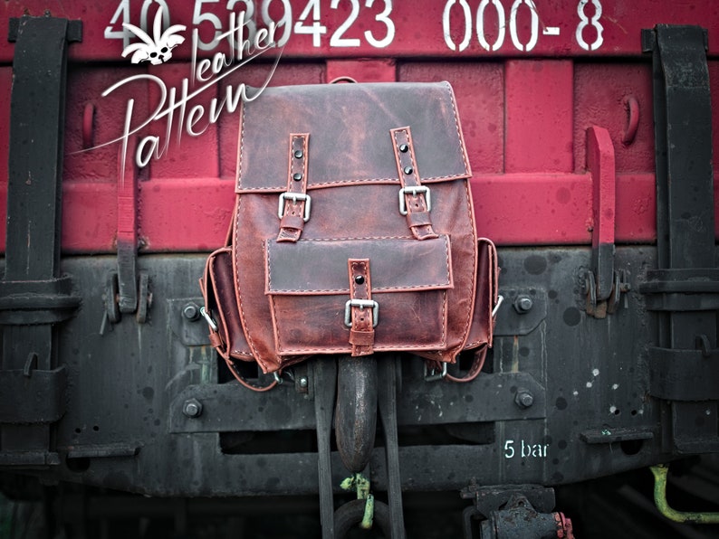 May include: A brown leather backpack with two large buckles and a smaller pocket on the front. The backpack is hanging from a metal hook on a red and black train car.