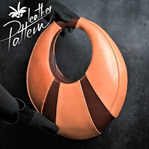 May include: A peach-coloured leather handbag with a brown leather accent. The bag has a unique, curved shape and a large, circular opening.