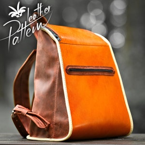 Puede incluir: Una mochila de cuero marrón y naranja con ribete blanco. La mochila tiene un bolsillo con cremallera en la parte posterior.