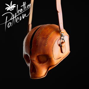 K&ouml;nnte beinhalten: Eine hellbraune Ledertasche in Form eines menschlichen Sch&auml;dels mit sichtbaren N&auml;hten, einem zentralen schwarzen Rei&szlig;verschluss und einem passenden Schulterriemen aus Leder. Der Text "Pattern Leather" steht wei&szlig; in der oberen linken Ecke.
