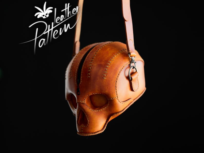 K&ouml;nnte beinhalten: Eine hellbraune Ledertasche in Form eines menschlichen Sch&auml;dels mit sichtbaren N&auml;hten, einem zentralen schwarzen Rei&szlig;verschluss und einem passenden Schulterriemen aus Leder. Der Text "Pattern Leather" steht wei&szlig; in der oberen linken Ecke.