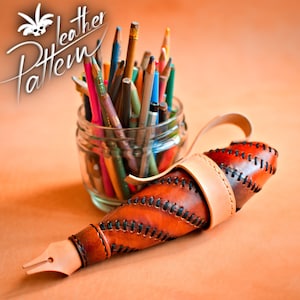 Può includere: Un portapenne in pelle marrone con un designo cucito e un cinturino. Il portapenne ha la forma di una punta di penna e si trova su una superficie di colore arancio chiaro. Un barattolo di vetro pieno di matite colorate si trova sullo sfondo.