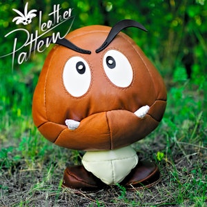 Puede incluir: Un juguete de peluche Goomba de cuero marrón con ojos y dientes blancos. El juguete está de pie sobre un lecho de hierba verde.