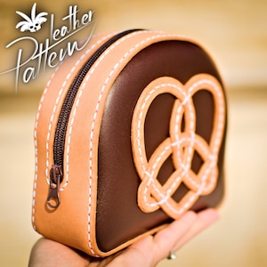 Puede incluir: Una bolsa de cuero marrón con cremallera, ribete de cuero beige y un diseño de pretzel cosido en blanco.