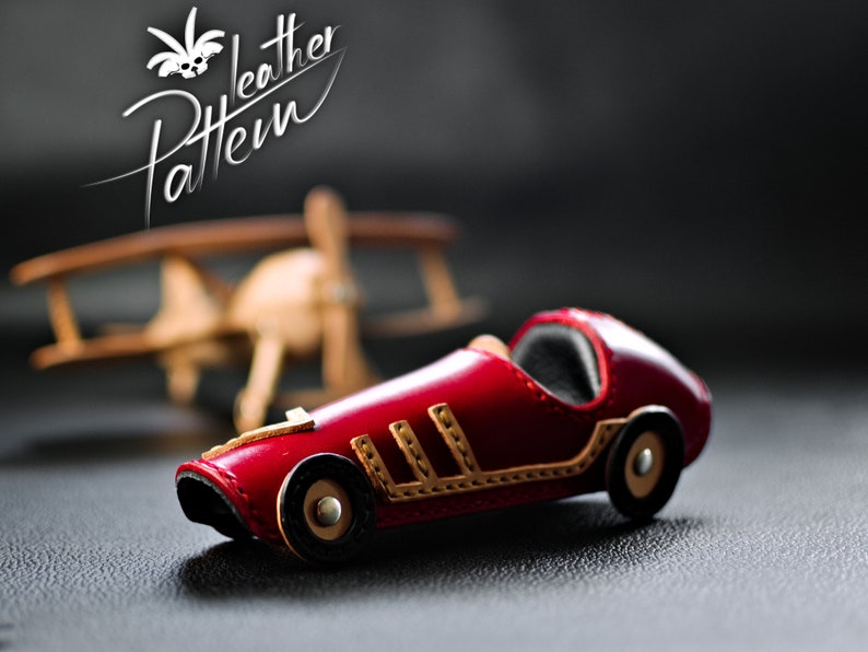 K&ouml;nnte beinhalten: Ein handgefertigtes rotes Leder-Rennwagen-Spielzeug mit beigen Akzenten und schwarzen R&auml;dern. Das Spielzeugauto hat ein klassisches Design und ist vor einem verschwommenen Hintergrund mit einem Holzflugzeug platziert. Das Bild enth&auml;lt den Text "Leather Pattern".