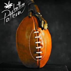 Pode incluir: Uma bola de futebol americano de couro marrom com costuras brancas. A bola está sendo segurada por uma mão robótica.