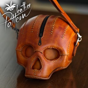 K&ouml;nnte beinhalten: Eine lohfarbene Ledertasche in Form eines menschlichen Sch&auml;dels mit einem schwarzen Rei&szlig;verschluss an der Oberseite. Die Tasche weist sichtbare N&auml;hte entlang der Kanten und Augenh&ouml;hlen auf. Der Text "Pattern Leather" ist oben links zu sehen.