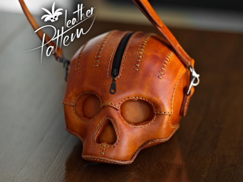 K&ouml;nnte beinhalten: Eine lohfarbene Ledertasche in Form eines menschlichen Sch&auml;dels mit einem schwarzen Rei&szlig;verschluss an der Oberseite. Die Tasche weist sichtbare N&auml;hte entlang der Kanten und Augenh&ouml;hlen auf. Der Text "Pattern Leather" ist oben links zu sehen.