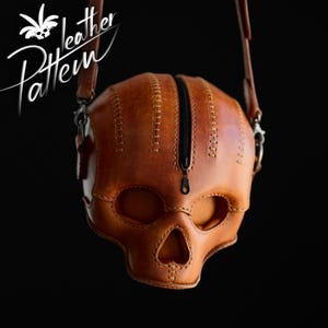 K&ouml;nnte beinhalten: Eine hellbraune Ledertasche in Form eines menschlichen Sch&auml;dels mit einem zentralen schwarzen Rei&szlig;verschluss, der von der Stirn bis zur Nase verl&auml;uft. Die Tasche hat sichtbare N&auml;hte und einen Schulterriemen aus Leder mit Metallbeschl&auml;gen.
