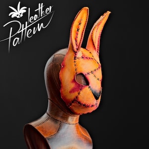 Può includere: Una maschera in pelle fatta a mano a forma di testa di coniglio, con lunghe orecchie e dettagli cuciti. La maschera è principalmente arancione con accenti neri e cuciture rosse. Le parole "leather Pattern" sono scritte in corsivo bianco.