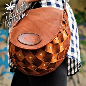May include: A brown leather saddle bag featuring a woven lattice pattern on the lower section, accented with small gold-tone metal rivets. The bag has a smooth flap with an oval patch embossed with the text Leatherhub and a shoulder strap.