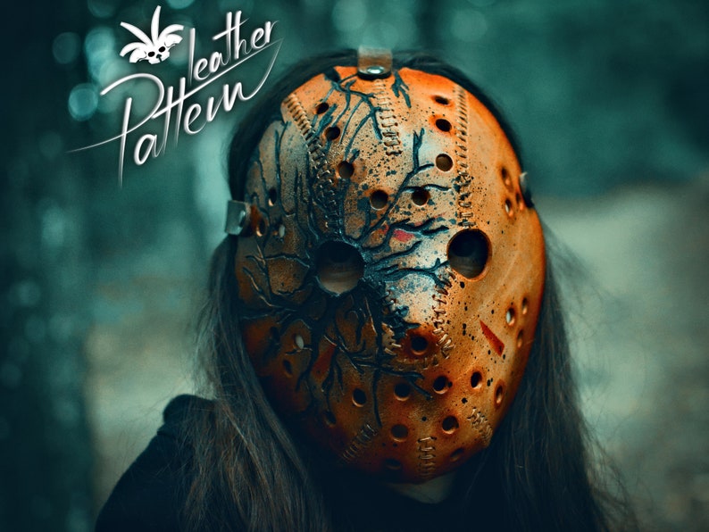 May include: A close-up of a brown leather hockey mask with black tree branches painted on it. The mask has a distressed look and is worn by a person with long dark hair.