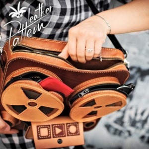 K&ouml;nnte beinhalten: Eine braune Ledertasche mit zwei runden F&auml;chern, die jeweils ein stilisiertes Design haben, das an eine Filmrolle erinnert. Die Tasche hat einen Rei&szlig;verschluss oben und einen verstellbaren schwarzen Riemen.