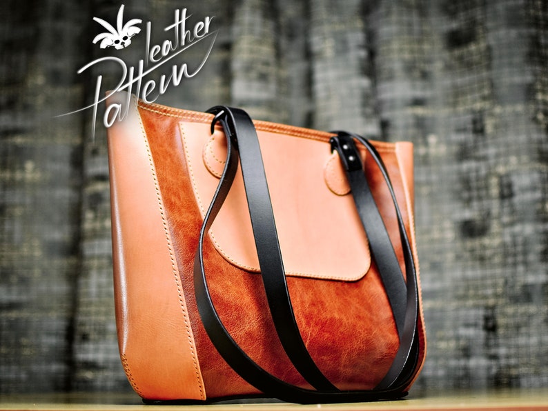 Puede incluir: Un bolso tote de cuero marr&oacute;n en dos tonos con un panel liso de color tostado claro y una secci&oacute;n texturizada de color marr&oacute;n oscuro. El bolso incluye dos correas largas de cuero negro sujetas con costuras visibles y remaches met&aacute;licos.
