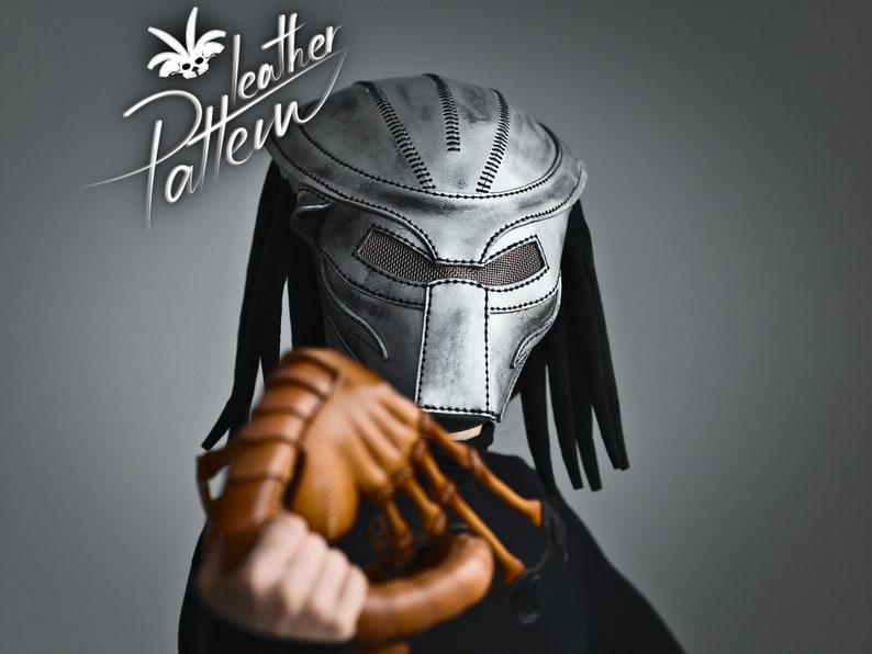 K&ouml;nnte beinhalten: Eine silberne Predator-Maske mit schwarzen Dreadlocks und einem braunen Lederhandschuh mit Krallen. Die Maske hat ein Netz-Augenabdeckung und ist aus Leder. Der Handschuh ist eine Requisite f&uuml;r ein Predator-Kost&uuml;m.