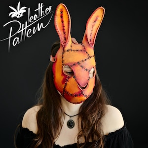 Puede incluir: Una máscara de conejo de cuero hecha a mano con tonos naranja y rojo, cosida con hilo negro. La máscara tiene orejas largas y erguidas y aberturas para los ojos. Las palabras "leather pattern" están escritas en blanco.