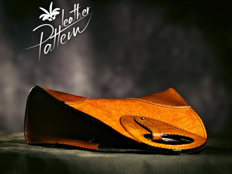 May include: A brown leather handbag with a unique design featuring a circular cutout and a buckle closure. The bag is lying on a dark surface.