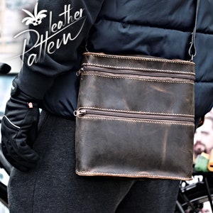 Puede incluir: Un bolso de cuero marrón cruzado con dos compartimentos con cremallera y una correa de cuero marrón. El bolso lo lleva una persona que lleva una chaqueta negra y pantalones grises.